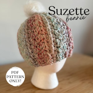 Puede incluir: Un gorro de ganchillo multicolor con un pompón blanco. El sombrero se muestra en una cabeza de maniquí blanca. El texto "Suzette beanie" está escrito en escritura negra. El texto "PDF PATTERN ONLY!" está en un círculo blanco.