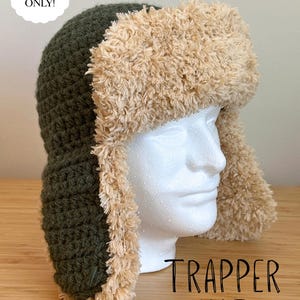 Puede incluir: Un gorro de trampero de ganchillo con corona y orejeras de color verde oliva oscuro, con un borde de piel sintética beige texturizada. La imagen incluye el texto PDF PATTERN ONLY! y TRAPPER HAT. El gorro se muestra sobre una cabeza de maniquí blanca.