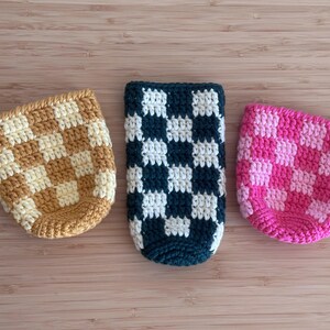 K&ouml;nnte beinhalten: Drei geh&auml;kelte Getr&auml;nkehalter mit Schachbrettmuster. Einer ist beige und cremefarben, ein weiterer marineblau und wei&szlig;, und der dritte rosa und hellrosa. Die Halter sind f&uuml;r Getr&auml;nkebeh&auml;lter in Standardgr&ouml;&szlig;e konzipiert.
