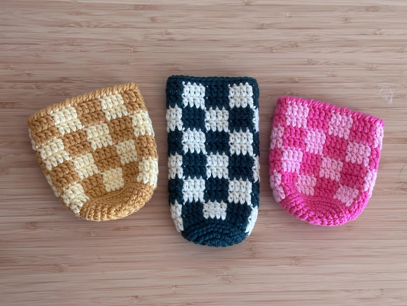 K&ouml;nnte beinhalten: Drei geh&auml;kelte Getr&auml;nkehalter mit Schachbrettmuster. Einer ist beige und cremefarben, ein weiterer marineblau und wei&szlig;, und der dritte rosa und hellrosa. Die Halter sind f&uuml;r Getr&auml;nkebeh&auml;lter in Standardgr&ouml;&szlig;e konzipiert.