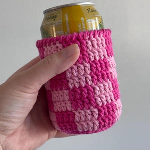K&ouml;nnte beinhalten: Ein handgefertigter Getr&auml;nkedosen-K&uuml;hler aus H&auml;kelarbeit in einem Schachbrettmuster aus hellem und dunklem Rosa. Der K&uuml;hler ist f&uuml;r eine Getr&auml;nkedose in Standardgr&ouml;&szlig;e konzipiert. Die oberen und unteren Kanten sind einfarbig dunkelrosa.