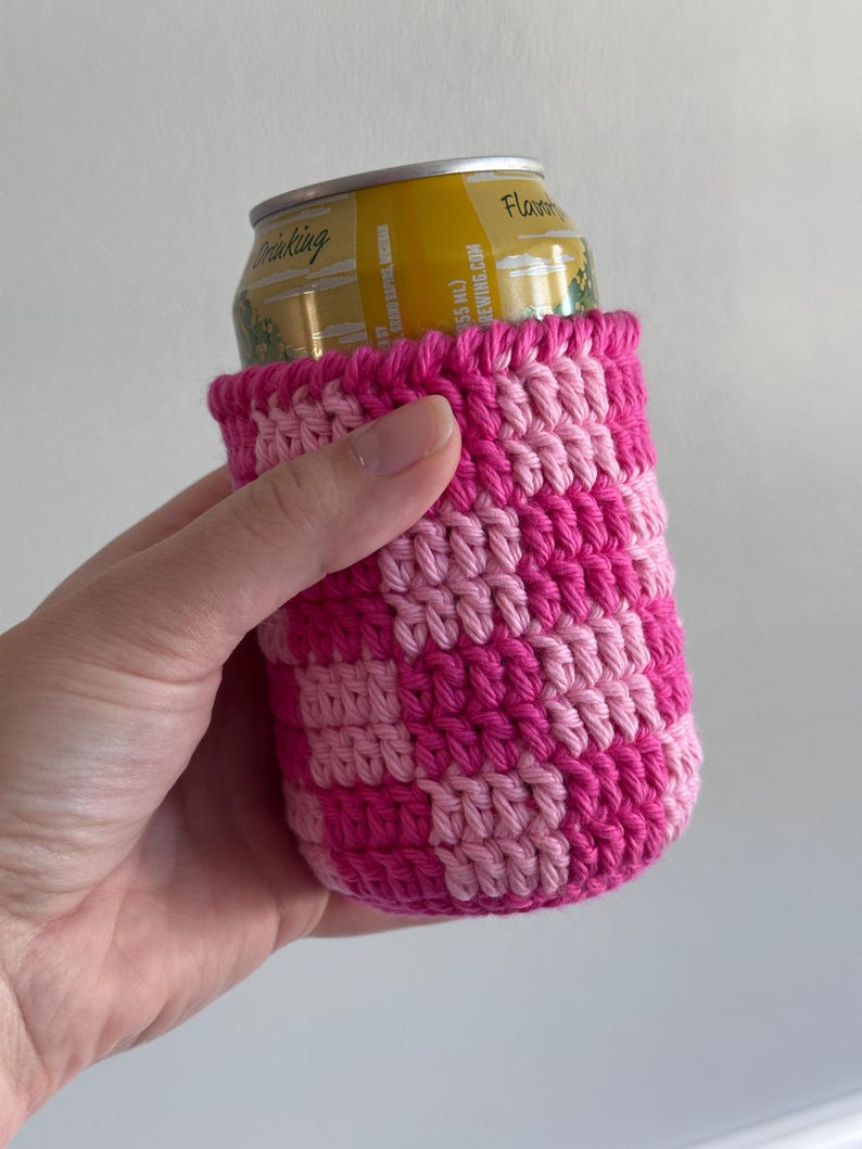 K&ouml;nnte beinhalten: Ein handgefertigter Getr&auml;nkedosen-K&uuml;hler aus H&auml;kelarbeit in einem Schachbrettmuster aus hellem und dunklem Rosa. Der K&uuml;hler ist f&uuml;r eine Getr&auml;nkedose in Standardgr&ouml;&szlig;e konzipiert. Die oberen und unteren Kanten sind einfarbig dunkelrosa.