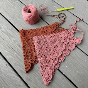 Op de afbeelding: Twee gehaakte driehoekige sjaals, een in een diepe roestkleur en de andere in een stoffige roze kleur, liggen op een houten oppervlak. Een bol stoffig roze garen en een haaknaald zijn ook zichtbaar.