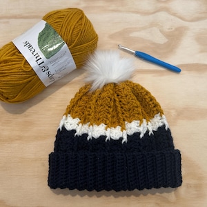Op de afbeelding: Een handgemaakte gehaakte muts in mosterdgeel, wit en marineblauw met een witte imitatiebont pompon. Een streng mosterdgeel garen en een blauwe haaknaald liggen naast de muts. Het garenlabel luidt "Lips & Threads".