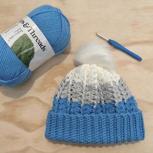 Op de afbeelding: Een blauwe, witte en grijze gehaakte muts met een witte pompon bovenop. De muts is gemaakt met een textuur-steek en heeft een blauwe bol wol en een haaknaald ernaast. Het etiket van de wolbol staat "Impeccable" en "Loops & Threads".