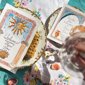 May include: Two watercolor save the date cards with a sun and moon design and a vase with flowers design. The cards are on a floral tablecloth with a glass of water and seashells.