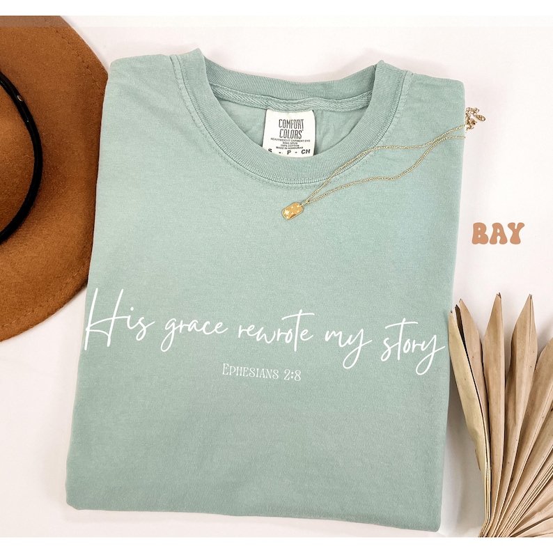 May include: A light green t-shirt with the text 'His grace rewrote my story' in white cursive font. The text 'Ephesians 2:8' is printed in white below the cursive text. The t-shirt is laying flat on a white surface.