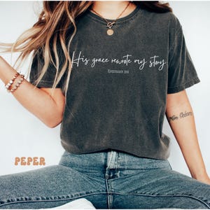 May include: A dark gray t-shirt with the text 'His grace rewrote my story' and the scripture reference 'Ephesians 2:8' in white text. The t-shirt is being worn by a person wearing blue jeans.