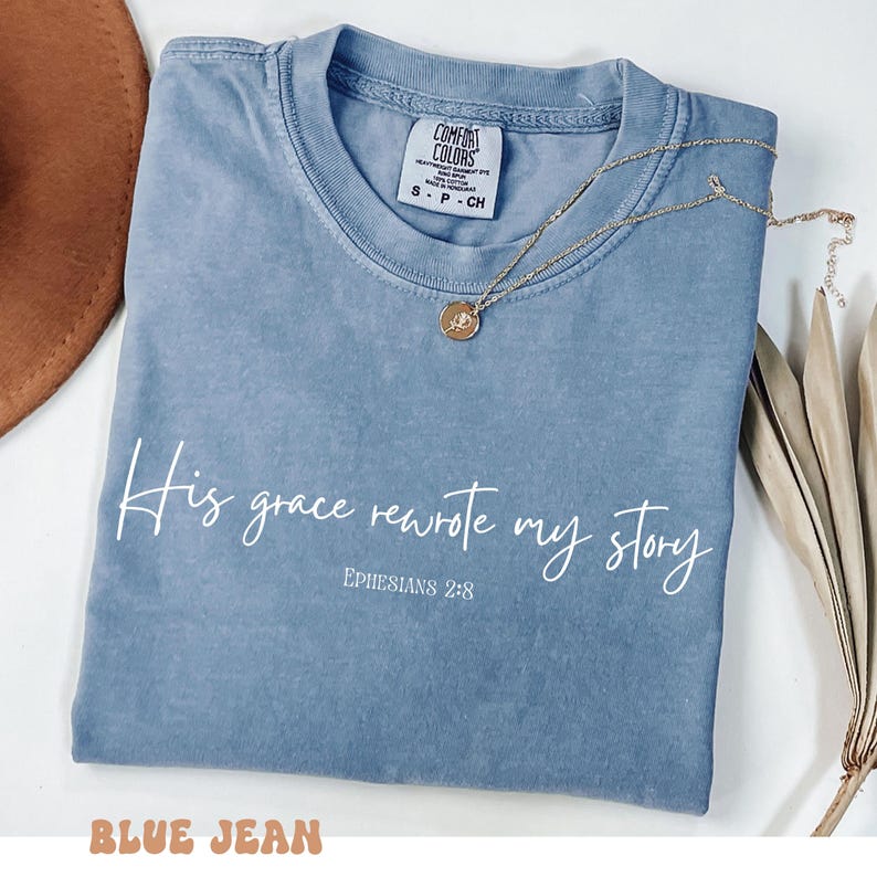 May include: A blue jean t-shirt with the text 'His grace rewrote my story' and 'EPHESIANS 2:8' in white. The shirt has a distressed look and is folded in half.