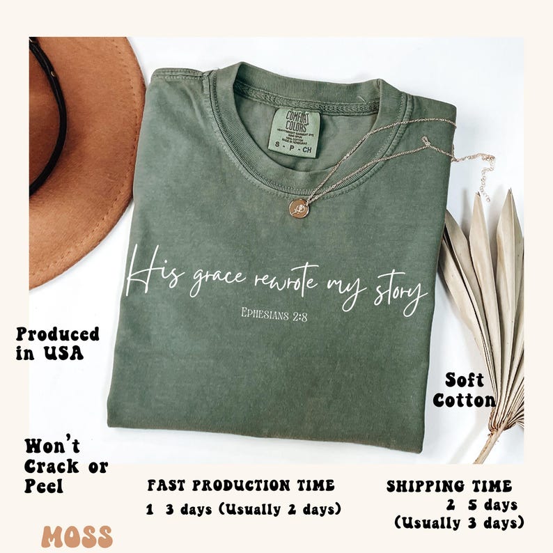 May include: A green t-shirt with the text 'His grace rewrote my story' and 'Ephesians 2:8' in white. The shirt is made of soft cotton and is produced in the USA. The shirt is displayed on a table with a brown hat and a dried plant.