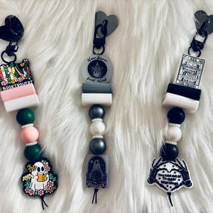 May include: Three black and white phone charms with different designs. The first charm features a stack of books with the text "Book Trovert" and a ghost holding a book. The second charm features a candle with the text "Slow Burn, Book Club". The third charm features a tarot card with the text "The Troves" and a skeleton hand with the text "You had me at "Trigger Warning"".