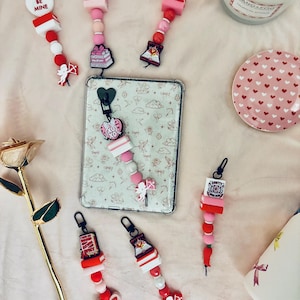 May include: Valentine's Day themed keychains with various charms, including hearts, books, and cupids. The keychains feature red, pink, and white beads, and some have text like "BE MINE" and "XOXO".