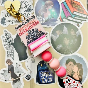 May include: A pink and white book charm with the text "Emotionally Attached to Fictional Characters". The charm is attached to a pink and white beaded keychain with a ghost charm.