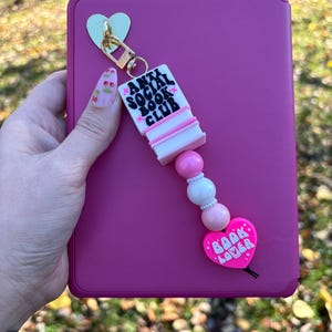 May include: A pink and white book charm with the text "Anti Social Book Club" and a pink heart charm with the text "Book Lover".