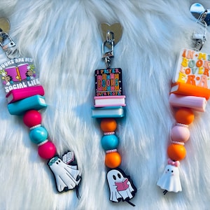 Könnte beinhalten: Drei bunte Schlüsselanhänger mit Buch-Motiv und Geisterfiguren. Die Anhänger zeigen die Sätze "Books are my social life", "I prefer books over people" und "In my book cover era".