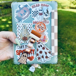 Könnte beinhalten: Eine transparente Handyhülle mit verschiedenen Halloween-Stickern, darunter Geister, Kürbisse und der Text "Read More Bookworms". Ein Perlenanhänger mit einem Geisteranhänger hängt an der Hülle.
