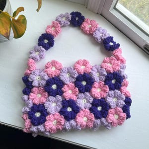 May include: A crocheted bag made of pink, purple, and lavender flowers. The bag is made of a series of crocheted flowers that are sewn together to form a bag.