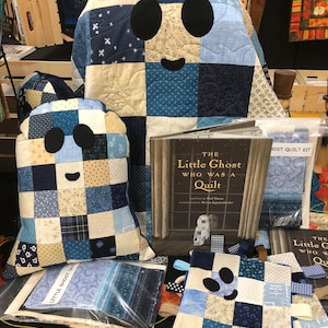 May include: A display of handmade ghost-shaped quilts and pillows in various shades of blue and cream. The ghost designs feature black oval eyes and a curved mouth. A book titled "The Little Ghost Who Was a Quilt" is also visible.