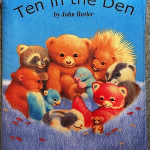 May include: A children's book cover titled "Ten in the Den" by John Butler. The cover features a blue background with illustrations of various cartoon animals, including bears, a raccoon, and a hedgehog. The animals are in a grassy area.