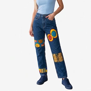 May include: Dark blue denim jeans featuring colorful fabric patches with circular and geometric patterns. The patches are sewn onto the thighs, knees, and lower legs of the pants. The jeans have a standard five-pocket design with a button fly closure.