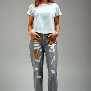 May include: A light blue t-shirt and distressed denim jeans with colorful patchwork pockets. The jeans have ripped details. The model is wearing dark blue sneakers with brown soles.