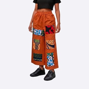 May include: Wide-leg, rust-colored corduroy pants with colorful patchwork designs. The patches feature various patterns and prints in shades of blue, red, green, and yellow. The pants have an elastic waistband and are worn with black sneakers.