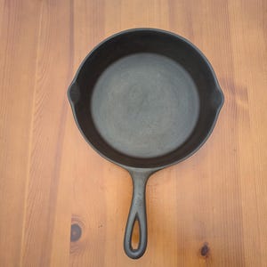 May include: A black cast iron skillet with a long handle and a rounded cooking surface. The skillet is shown from a top-down perspective, resting on a light brown wooden surface. The handle has a hole for hanging.