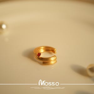 May include: A gold ring with a red stone set in the center. The ring is made of multiple bands that are twisted together.