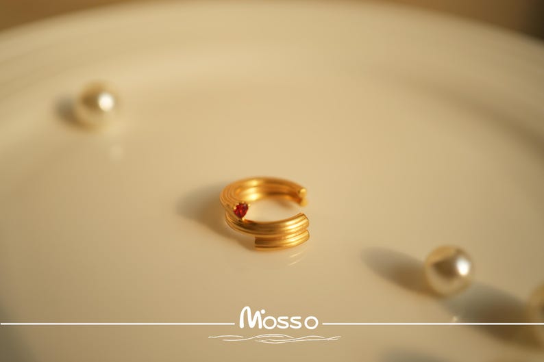 May include: A gold ring with a red stone set in the center. The ring is made of multiple bands that are twisted together.