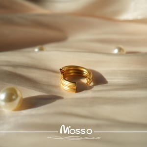 May include: A gold ring with a red gemstone on a white fabric background. The ring is open and has a textured design.