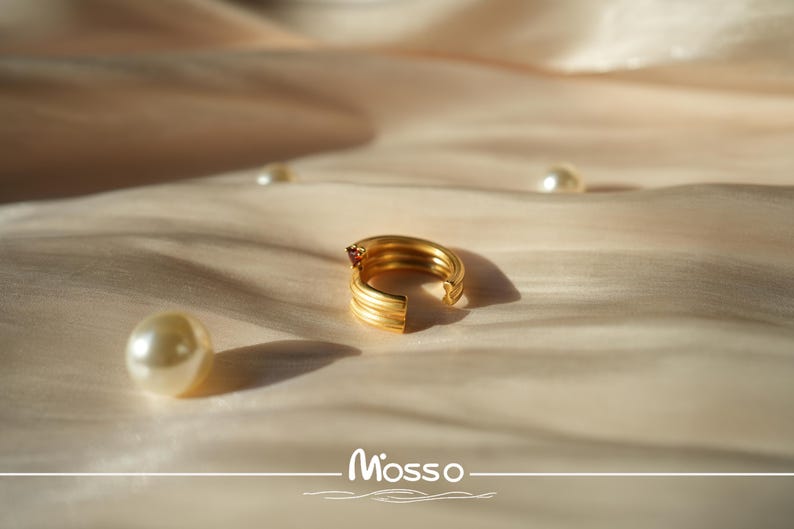 May include: A gold ring with a red gemstone on a white fabric background. The ring is open and has a textured design.