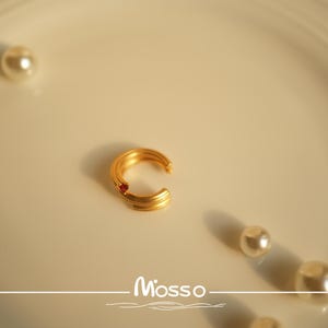 May include: A gold-toned ear cuff with a single red gemstone. The cuff is on a white surface with the word "Mosso" written in a decorative font.