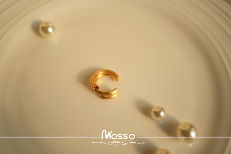 May include: A gold-toned ear cuff with a single red gemstone. The cuff is on a white surface with the word "Mosso" written in a decorative font.