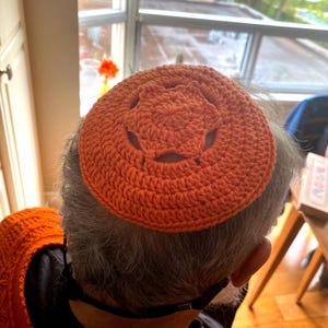 May include: A crocheted kippah in a bright orange color with a star-shaped design. The kippah is worn on top of a person's head.