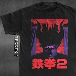 May include: A black short-sleeved t-shirt featuring a graphic print of a character with spiky hair and red eyes against a red and purple background. The text TEKKEN 2 is printed vertically on the left, and the Japanese characters 鉄拳2 are below.