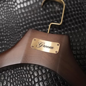 May include: A dark brown wooden clothes hanger featuring a polished gold-tone metal hook and a rectangular brass-colored nameplate centered on the front. The nameplate is secured with four small screws and has the word Groom engraved in black script.