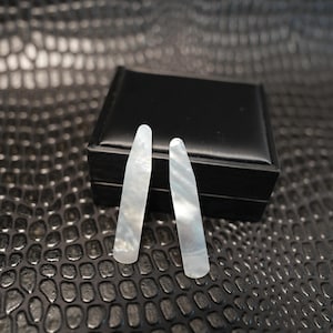 May include: A pair of white mother-of-pearl collar stays with a smooth, iridescent finish and a tapered, elongated shape. These accessories feature a rounded bottom edge and a pointed top, showcasing the natural luster of the shell material.
