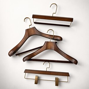 May include: A collection of four dark brown wooden clothes hangers with brass-toned metal hooks. Two hangers feature wide, contoured shoulders, one includes a horizontal bar with a dark velvet grip, and one has a bar with two adjustable clips.