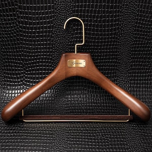 May include: A dark brown wooden clothes hanger featuring a gold-toned metal hook and a matching gold-toned rectangular plaque centered on the front. The plaque is engraved with the word Groom in a cursive script. A brown velvet-covered bar is included.