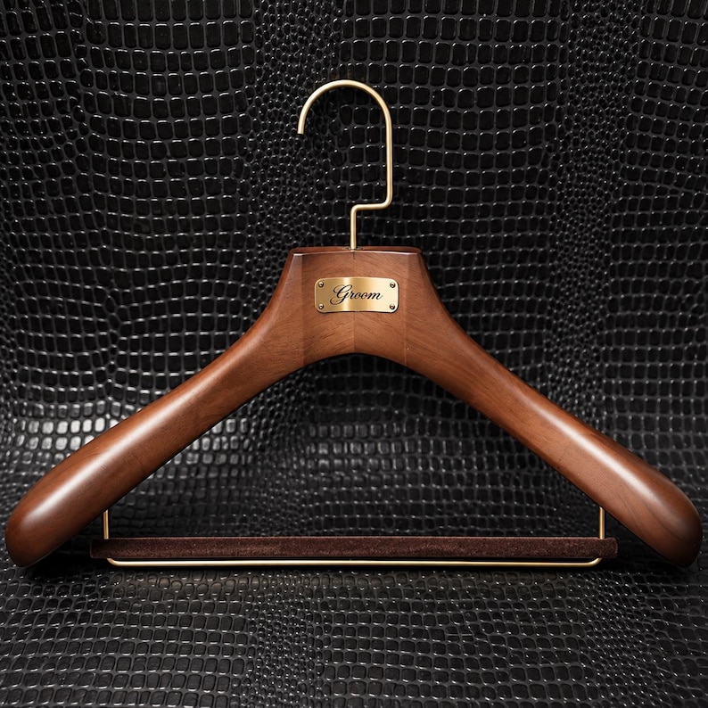 May include: A dark brown wooden clothes hanger featuring a gold-toned metal hook and a matching gold-toned rectangular plaque centered on the front. The plaque is engraved with the word Groom in a cursive script. A brown velvet-covered bar is included.
