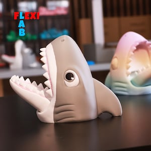 May include: A gray shark-shaped figurine with an open mouth revealing white teeth and a pink interior. The shark has a large eye and fins. The word "FLEXI AB" is visible in the background. Another shark figurine is visible in the background.