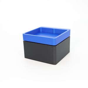 May include: A two-piece container with a square shape. The base is black, and the lid is a vibrant blue. The lid has a recessed interior, creating a tray-like design. The container is likely made of plastic.
