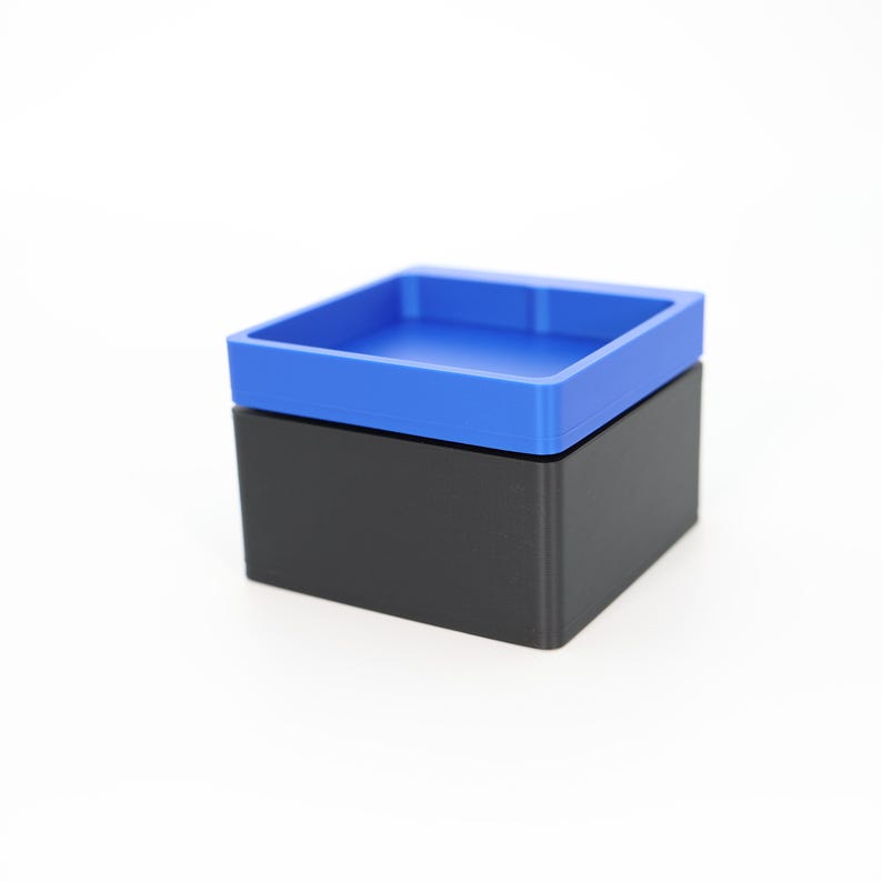 May include: A two-piece container with a square shape. The base is black, and the lid is a vibrant blue. The lid has a recessed interior, creating a tray-like design. The container is likely made of plastic.