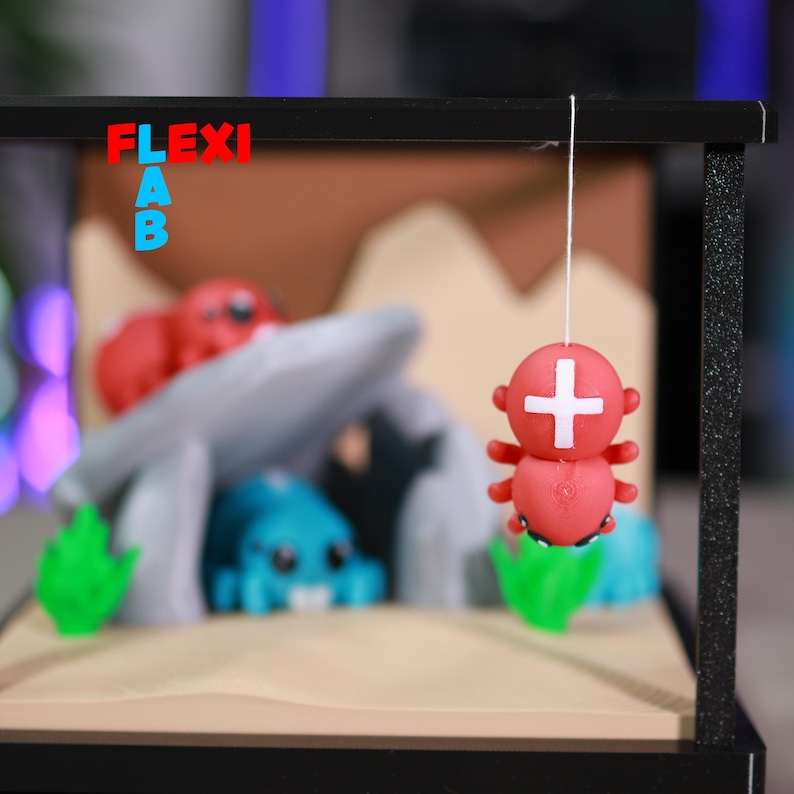 May include: A miniature diorama featuring a red and white cross-shaped figure suspended by a string. The scene includes a cave, blue and red figures, and green plant-like shapes. The word "FLEXI LAB" is displayed in red and blue.