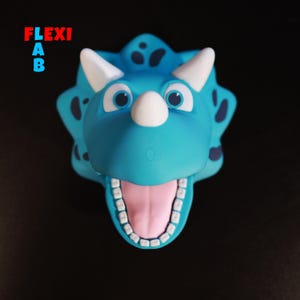 Peut inclure: Une marionnette de dinosaure bleue et blanche avec une grande bouche ouverte et une langue rose. Le dinosaure a une corne blanche et de grands yeux.