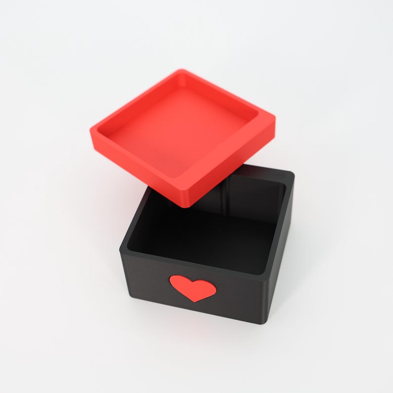 K&ouml;nnte beinhalten: Eine schwarze quadratische Box mit rotem Deckel und einem roten Herz auf der Vorderseite. Der Deckel ist leicht ge&ouml;ffnet und gibt den Blick auf das schwarze Innere der Box frei. Die Box wird wahrscheinlich zur Aufbewahrung oder als Dekorationsartikel verwendet.
