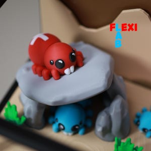 May include: A red spider figurine with a white cross on its back, perched on a gray rock formation. Below, a blue spider figurine is visible. The word "FLEXI AB" is written in red and blue.