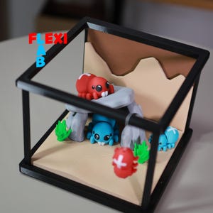 May include: A miniature diorama featuring several colorful spider-like creatures within a black framed enclosure. The spiders are red, blue, and red with white accents. The background includes a tan landscape with rock formations and green foliage.