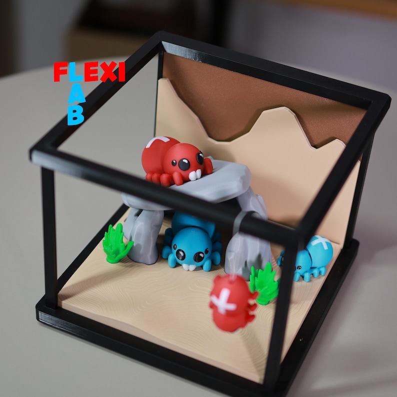 May include: A miniature diorama featuring several colorful spider-like creatures within a black framed enclosure. The spiders are red, blue, and red with white accents. The background includes a tan landscape with rock formations and green foliage.