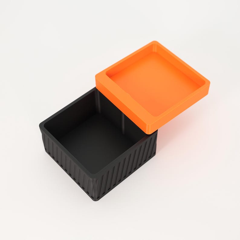 Puede incluir: Un recipiente cuadrado negro con un exterior texturizado y acanalado y una tapa naranja abierta. La tapa es cuadrada y se encuentra ligeramente por encima del recipiente. El recipiente est&aacute; vac&iacute;o y se encuentra sobre una superficie blanca.